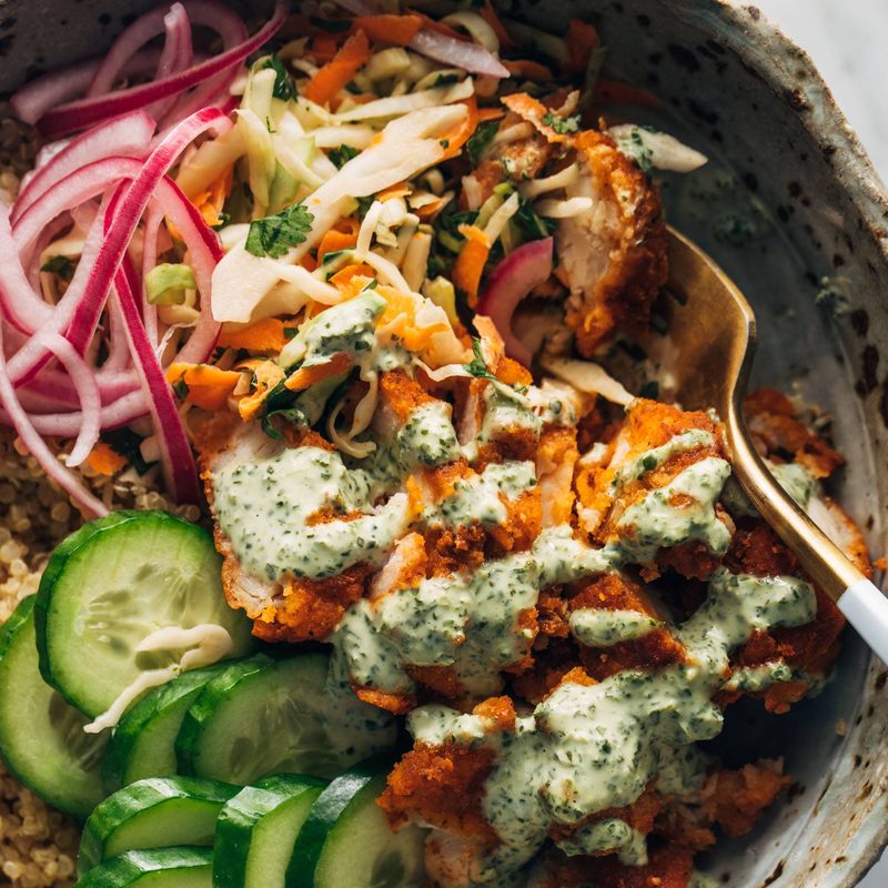 Crispy chicken, cucumbers, quinoa, and pickled red onions in a bowl with mint sauce and a fork